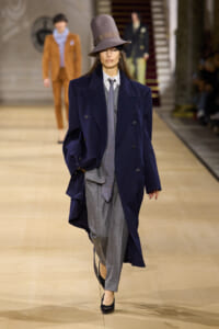 Model walking a fashion runway in an oversized navy double-breasted coat over a gray suit and tie, with a tall floppy gray hat.