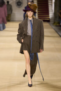 Model walks the runway in a brown oversized tweed blazer and matching skirt, checkered shirt, blue tie, and wide brim hat.