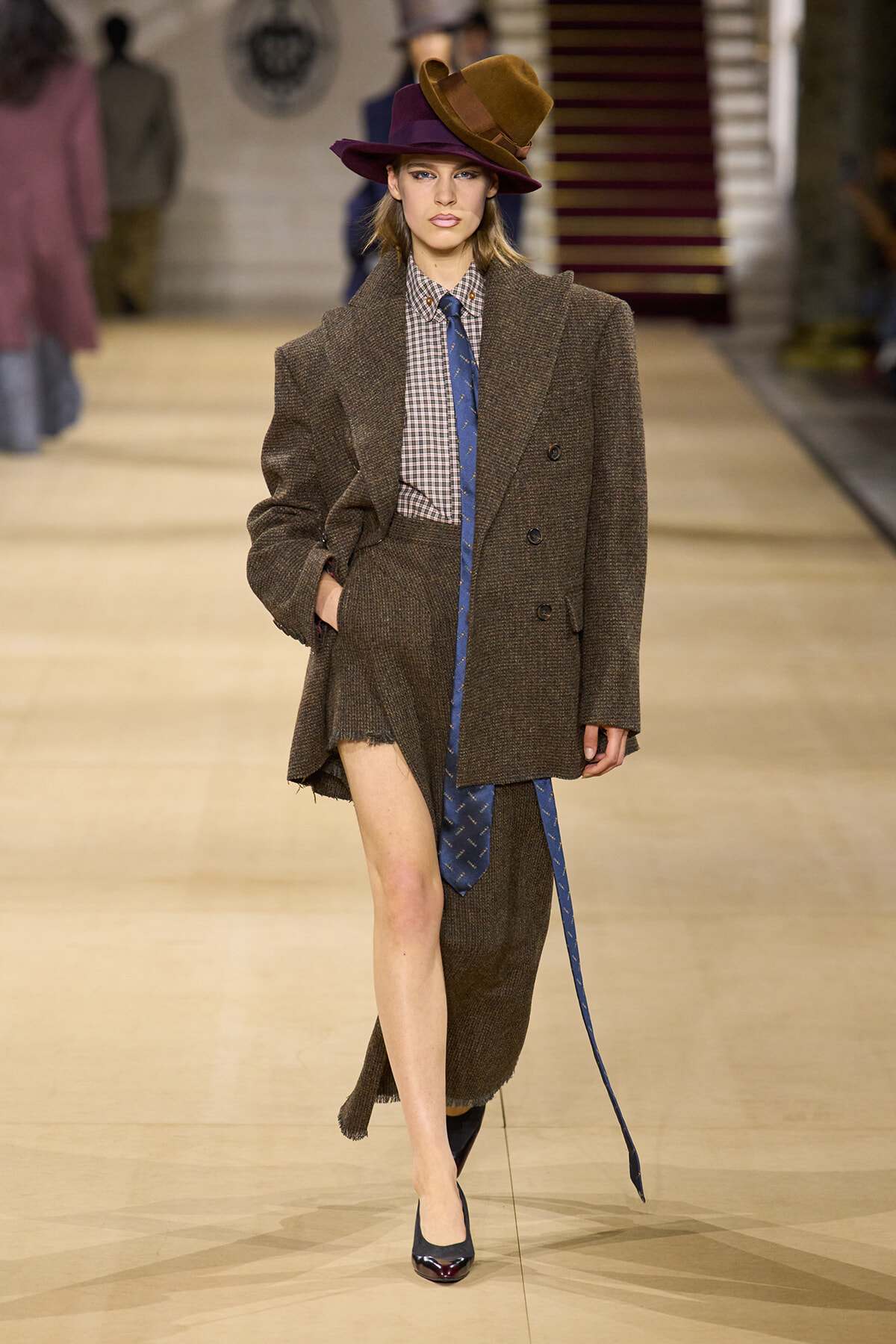 Model walks the runway in a brown oversized tweed blazer and matching skirt, checkered shirt, blue tie, and wide brim hat.