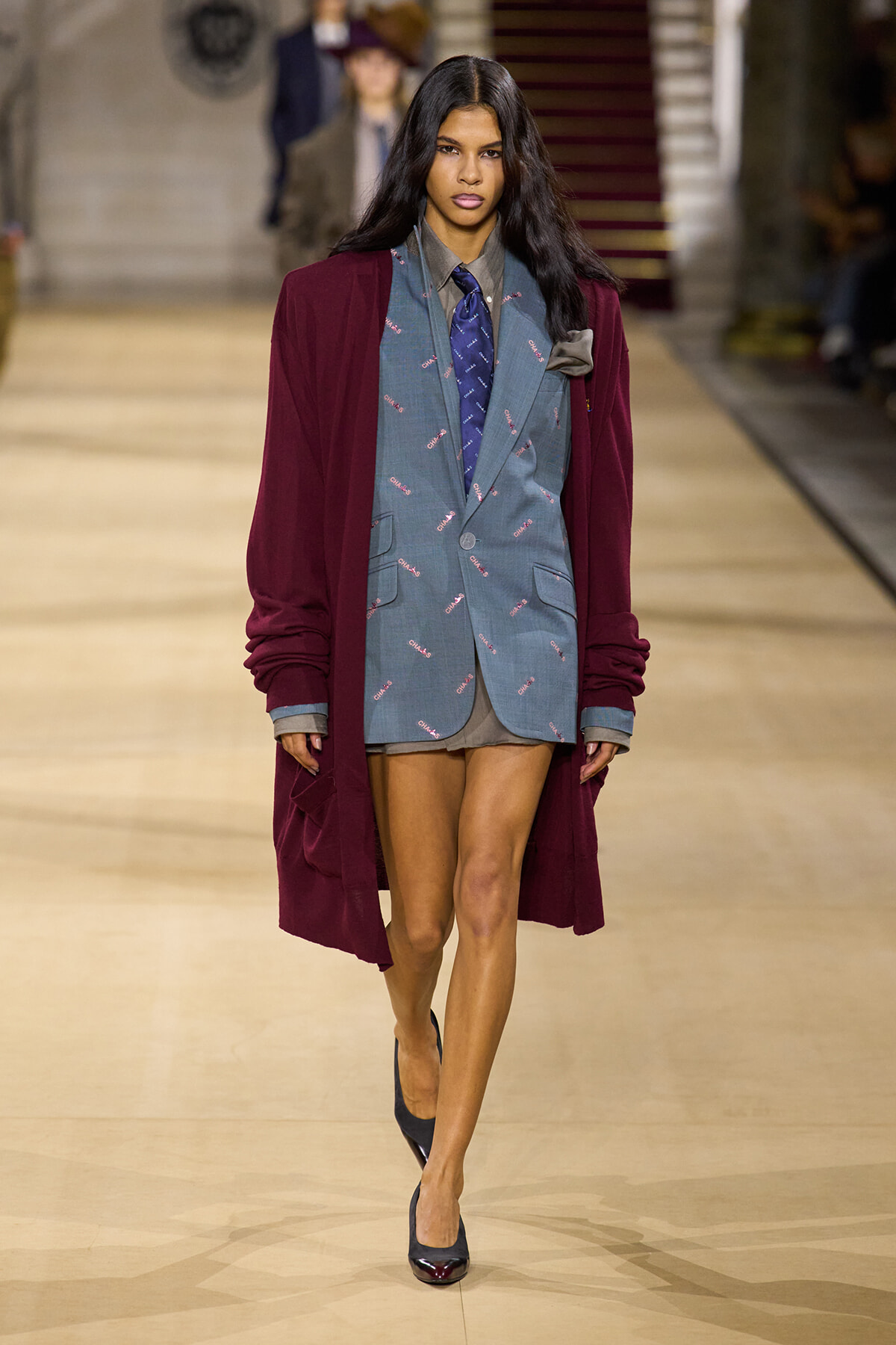 Model walks a runway wearing a burgundy long coat over a blue patterned blazer, grey shirt, blue tie, and dark heels.