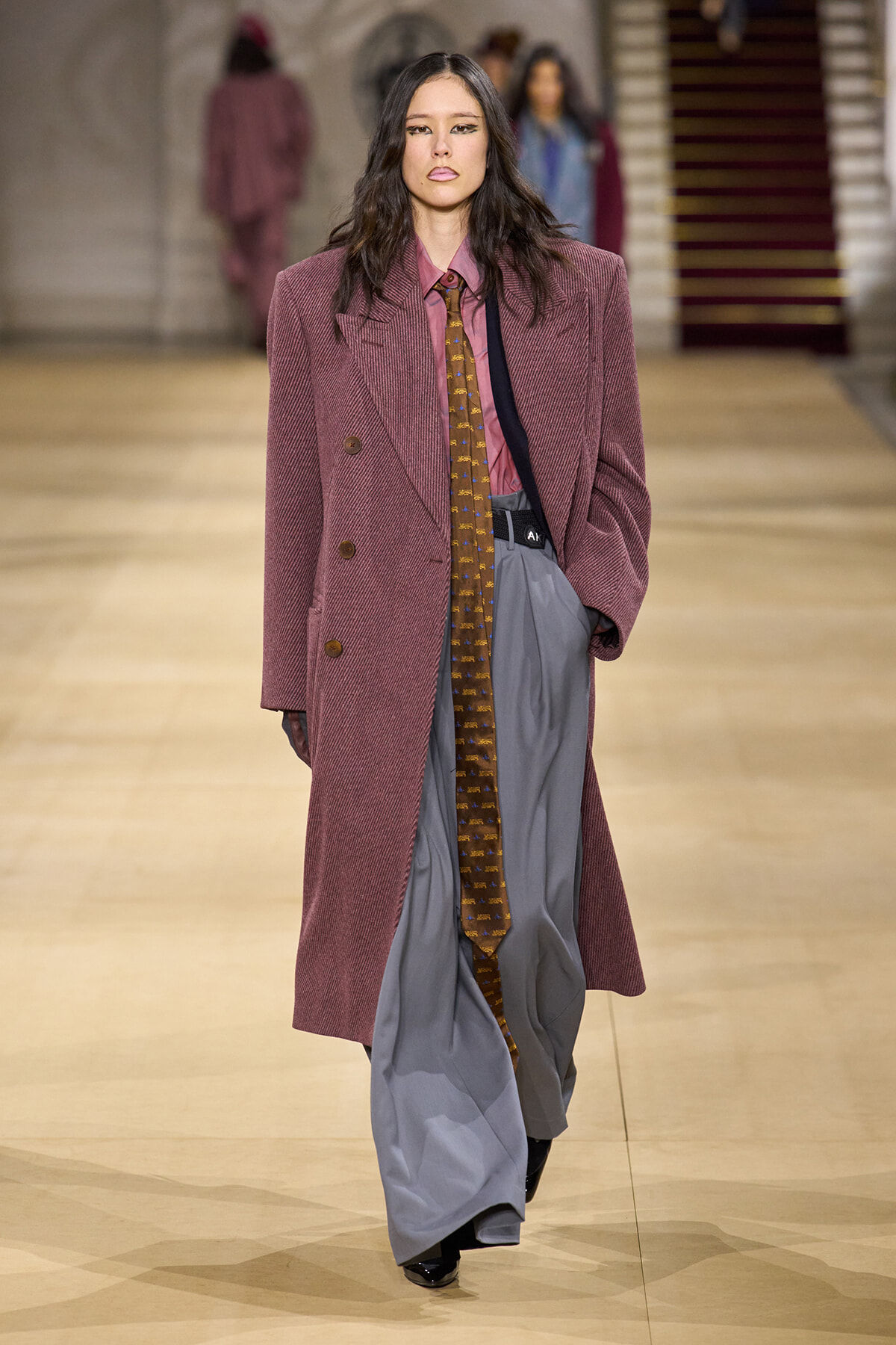 Model walks the runway in an oversized mauve blazer over a pink shirt, with a long patterned tie and loose gray wide-leg trousers.