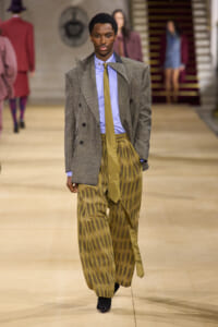 Male model on a runway wearing a gray textured blazer, light blue shirt, mustard tie, and oversized gold-striped wide-leg pants.