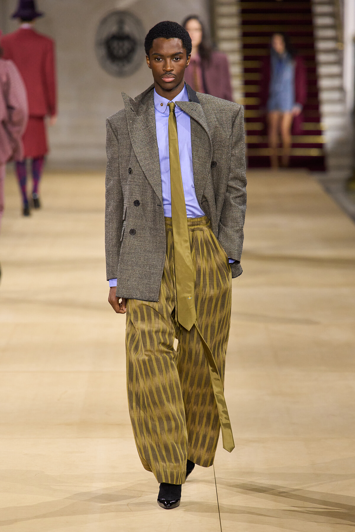 Male model on a runway wearing a gray textured blazer, light blue shirt, mustard tie, and oversized gold-striped wide-leg pants.