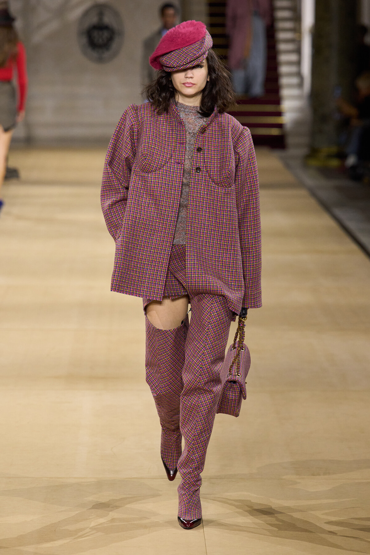 Model on a fashion runway wearing a coordinated pink plaid suit—oversized coat and matching pants with a knee opening—paired with burgundy heels and a pink beret. She carries a small pink bag.