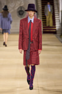 Male model on a fashion runway wearing a red checkered suit with a blue shirt, dark tie, and purple hat, plus mismatch blue-red patterned tights.