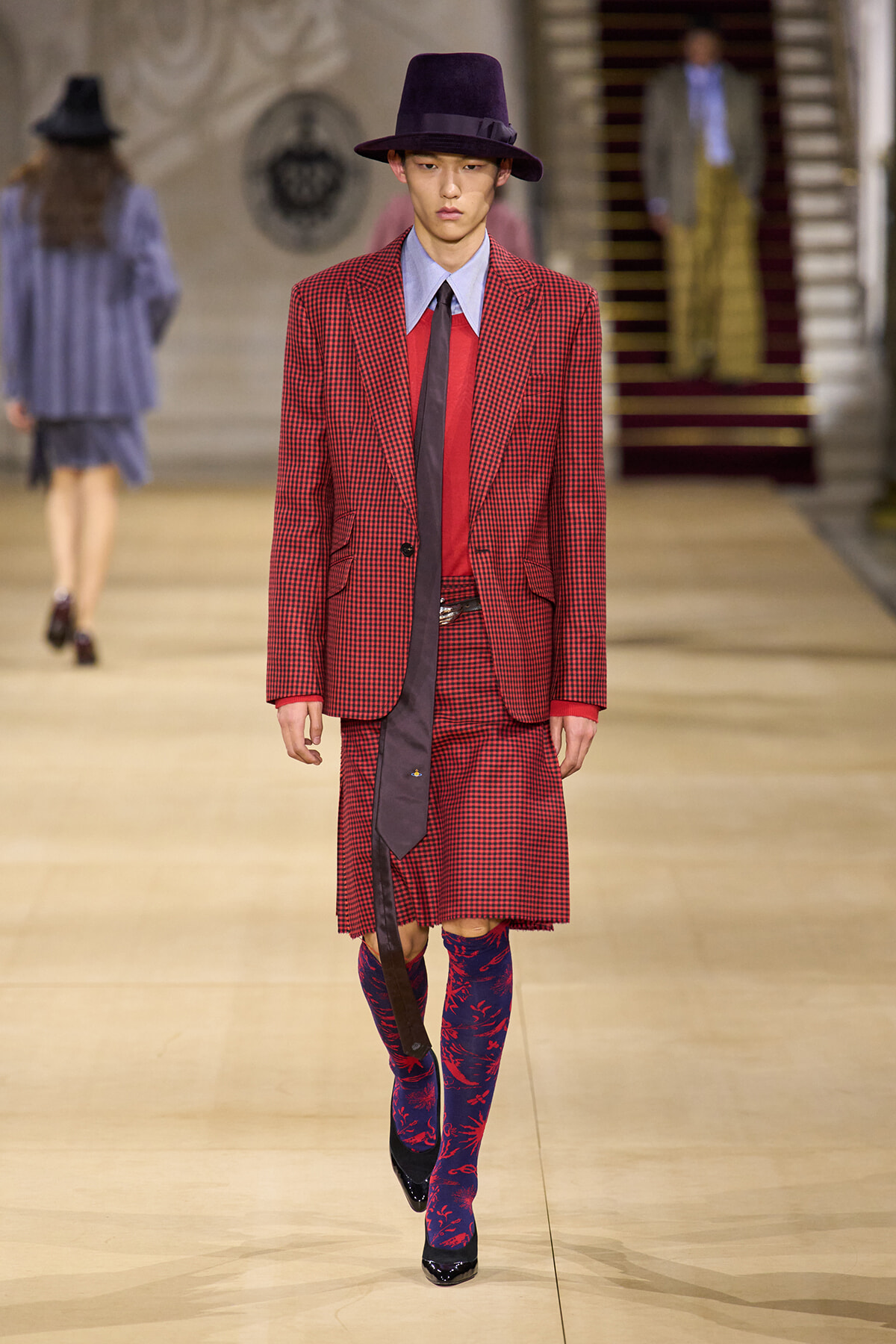 Male model on a fashion runway wearing a red checkered suit with a blue shirt, dark tie, and purple hat, plus mismatch blue-red patterned tights.