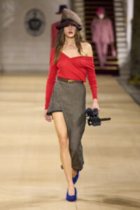 Model on a runway wearing a red off-shoulder top, gray wrap skirt, hat, and blue shoes while carrying a professional video camera.