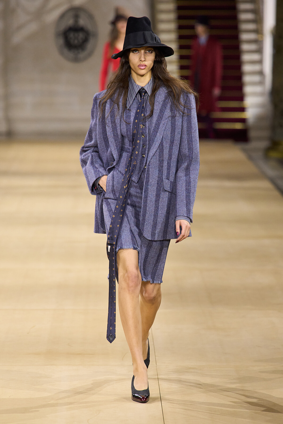 Model walks the runway in a blue striped oversized suit with matching shorts, striped shirt, navy tie, black wide-brim hat, and black heels.