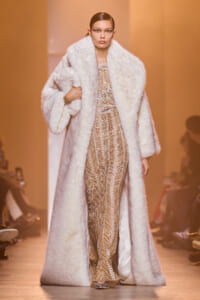 Model on a runway wearing a pale fur coat over a gold sequined gown, walking down the catwalk.