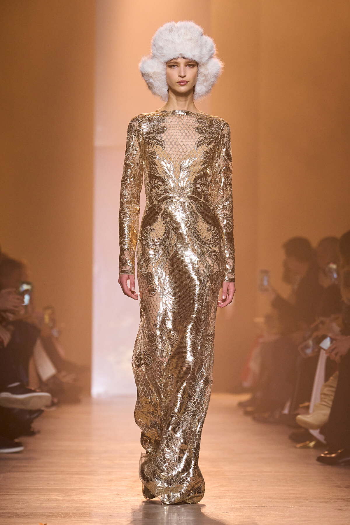 Model walks the runway in a full-length gold sequined lace gown with a sheer neckline and a white fluffy fur hat.