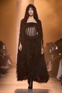Model walks the runway in a long, black feathered hooded gown with a sheer striped corset bodice and black ankle boots.