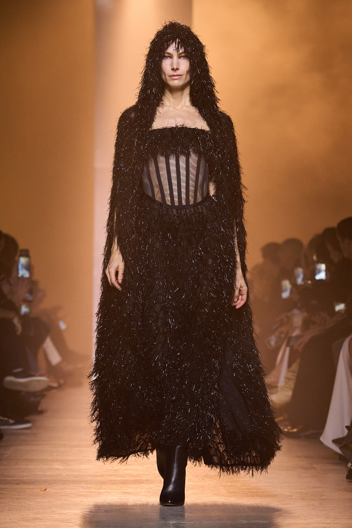 Model walks the runway in a long, black feathered hooded gown with a sheer striped corset bodice and black ankle boots.