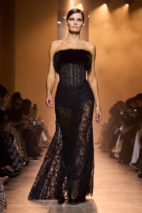 Model walking a runway in a strapless black lace gown with a fur bust, spotlighted against a warm background.