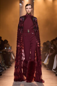 Model walks the runway in a burgundy jumpsuit with a jeweled cape and tall fur boots.