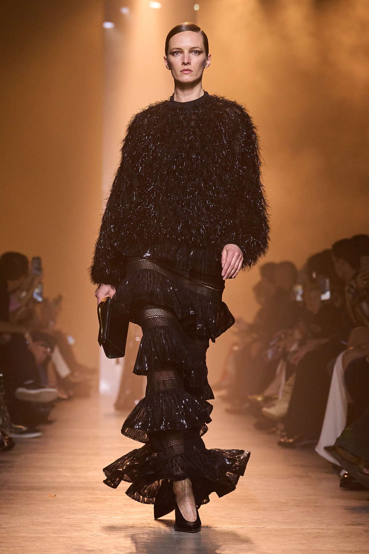 Model on a runway wearing a black textured feathered sweater with a multi-tiered black ruffled skirt, carrying a black clutch as she walks forward in dramatic lighting.