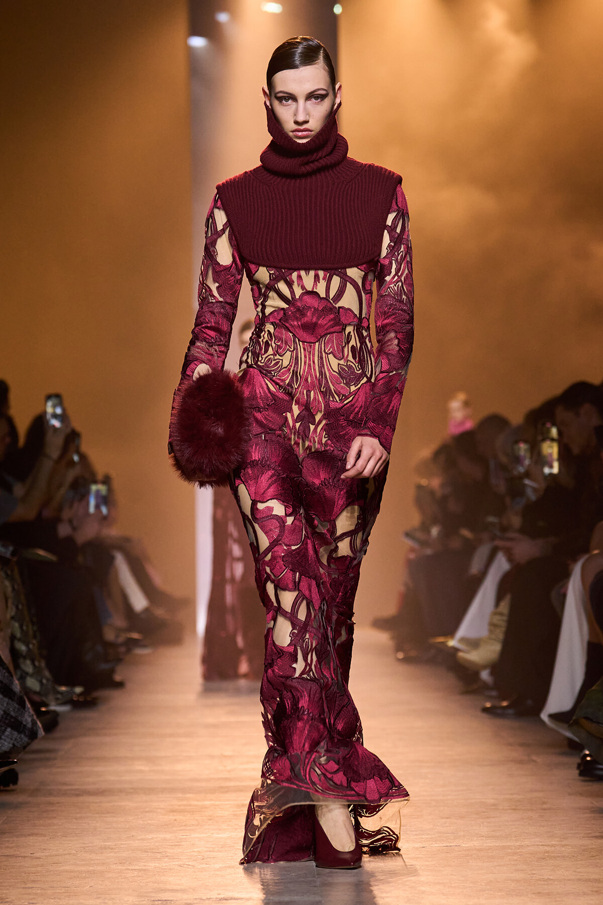 Runway model in a burgundy lace gown with a chunky knit burgundy turtleneck, carrying a burgundy fur clutch.