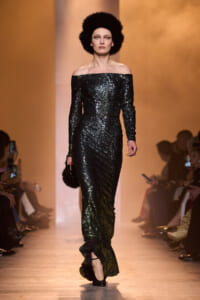 Model on a runway wearing an off-shoulder, floor-length black sequin gown with a large black fur hat; she carries a small fur clutch against an warm orange backdrop.