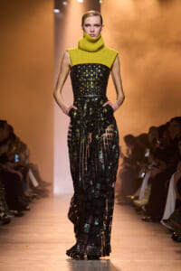 Model on a runway wearing a lime-yellow chunky knit turtleneck over a black mosaic-sequin gown, hands in pockets.