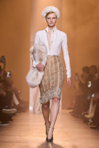 Model walking a runway in a white zip-front blouse, gold beaded knee-length skirt, and white fur hat with a matching fur stole.