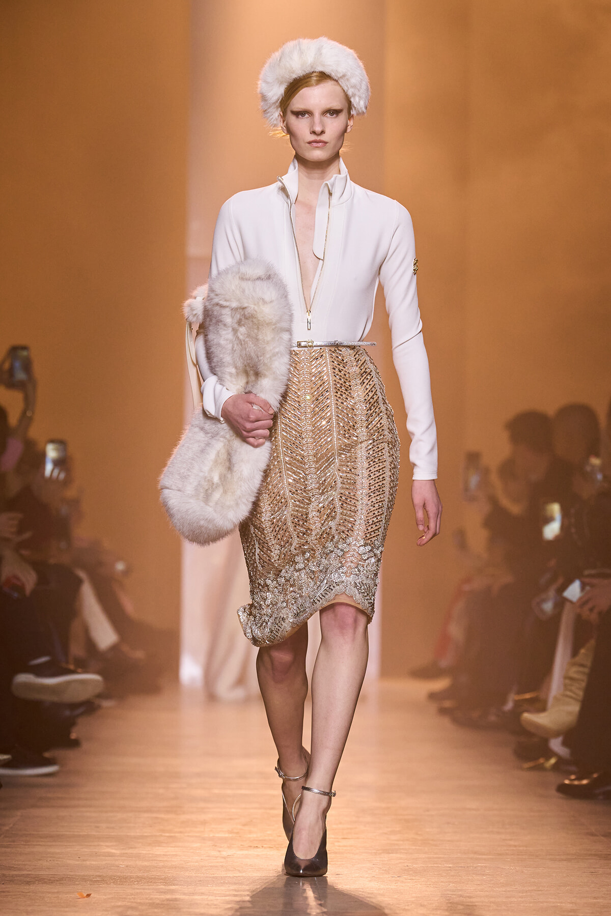 Model walking a runway in a white zip-front blouse, gold beaded knee-length skirt, and white fur hat with a matching fur stole.