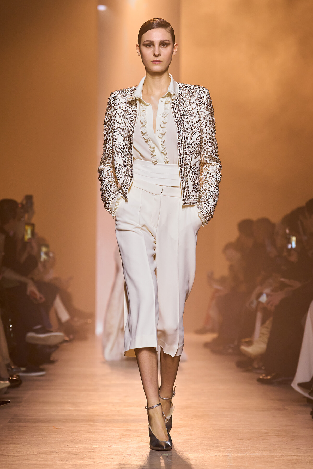 Model walks the runway in an ivory satin suit and a rhinestone-embellished jacket.