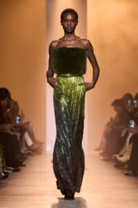 Model walking the runway in a strapless green furry bandeau and shimmering green-to-black pleated pants, hands in pockets, audience blurred in background.