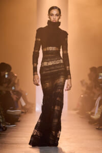 Model on a runway wearing a long black lace dress with a high turtleneck and sheer panels.