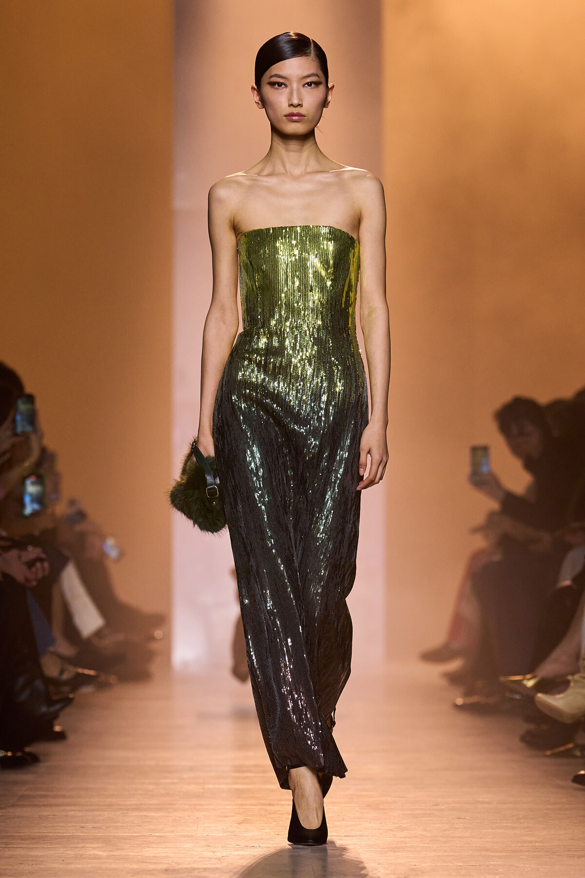 Model walks the runway wearing a strapless sequined gown that fades from emerald to black, holding a small fur-accented clutch on her left.