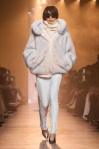 Model walks the runway in a oversized pale fur coat, cream turtleneck, white trousers, and dark sunglasses; spotlighted catwalk scene.