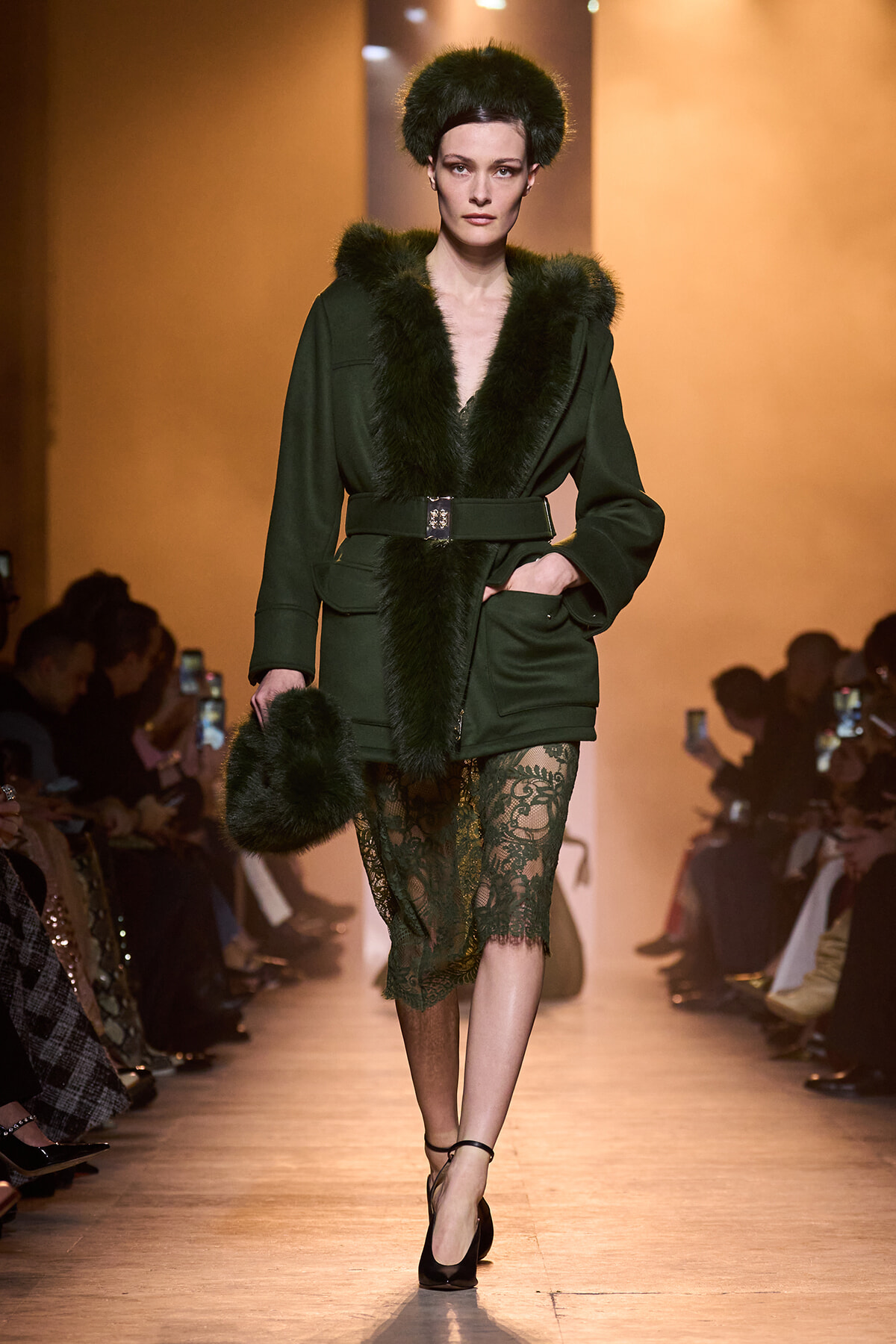 Model walking a runway in a dark green fur-trimmed coat with a matching fur hat, carrying a fur bag and wearing lace dress underneath.