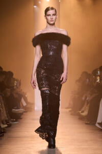 Model walks the runway in a black sequined off-shoulder gown with fur trim and ankle boots.