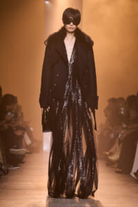 Runway model in a black fur-collared coat over a sheer, sequined gown with oversized sunglasses and a small black clutch.