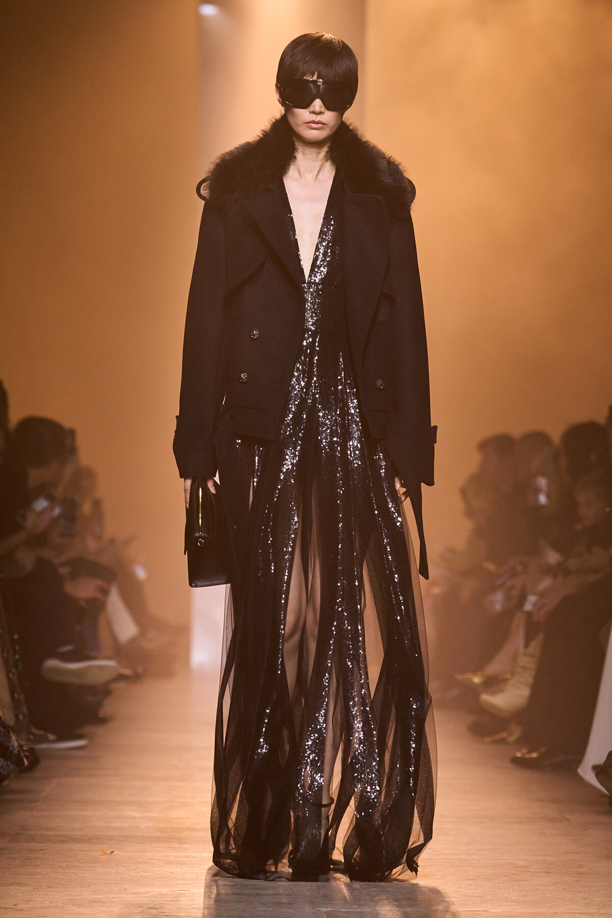 Runway model in a black fur-collared coat over a sheer, sequined gown with oversized sunglasses and a small black clutch.