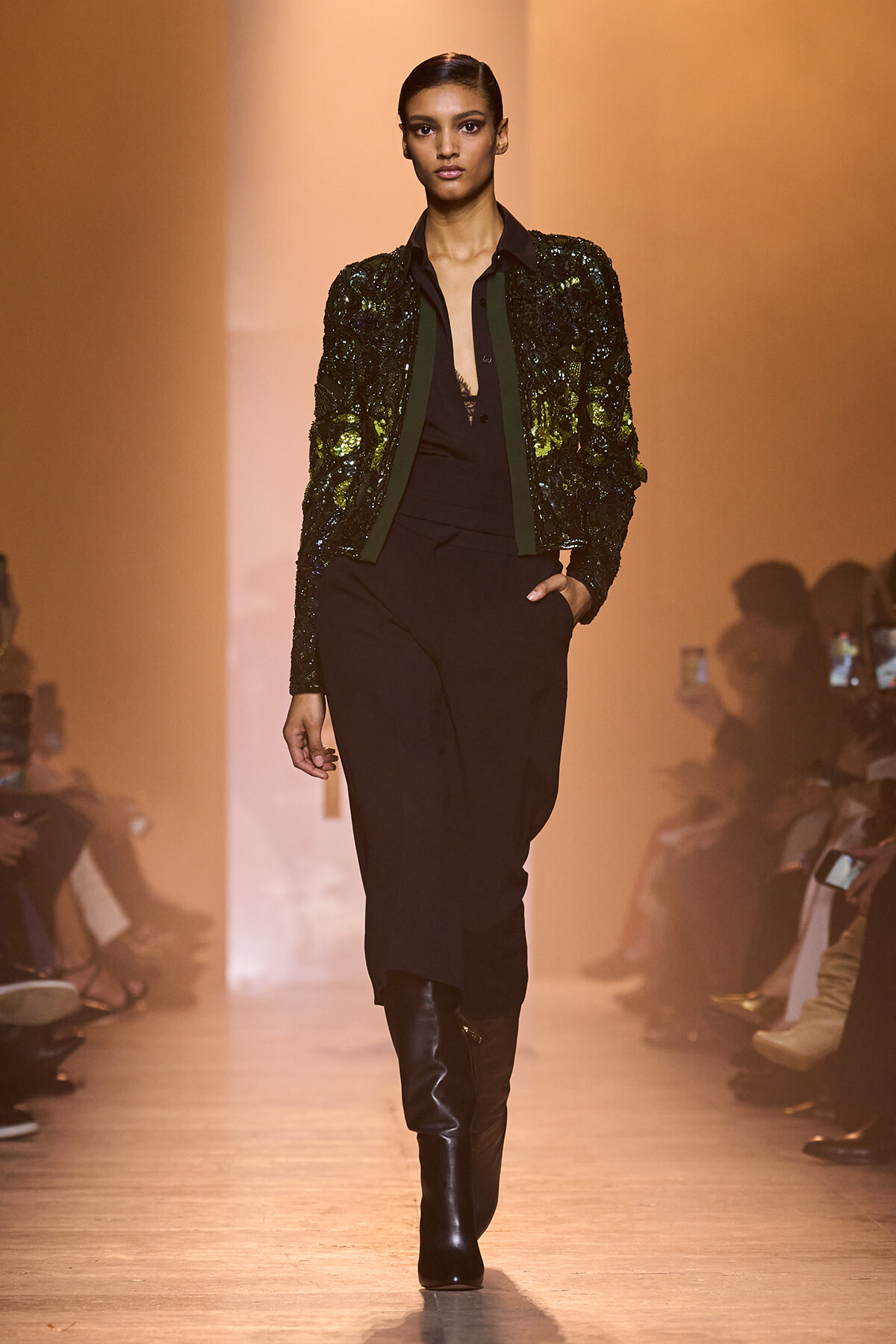 Model walking the runway in a black outfit with a dark green sequined jacket and knee-high black boots.