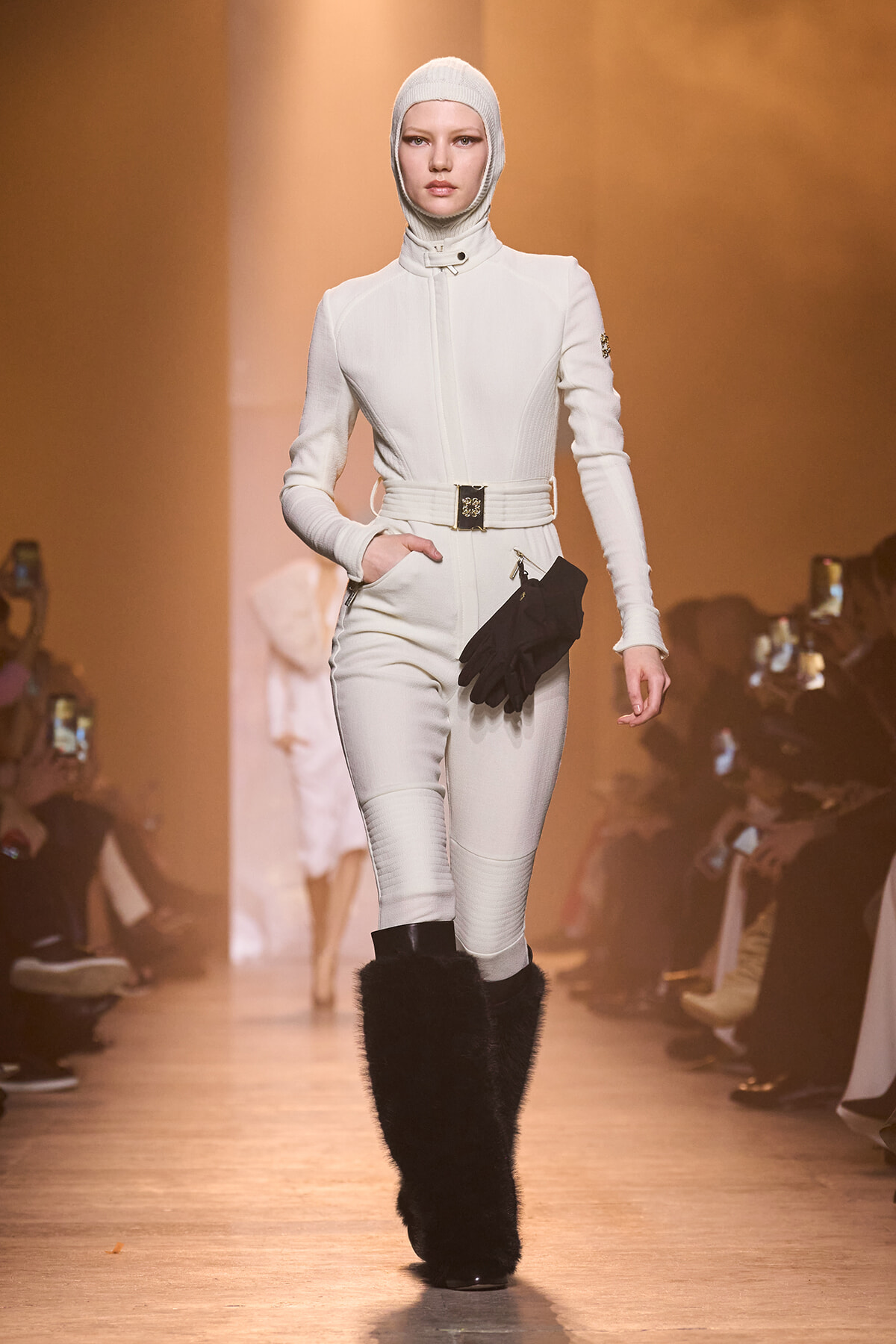 Model walks a fashion runway in an all-white, tailored jumpsuit with a matching knit hood, belt, and knee-high black fur boots, holding gloves in one hand.