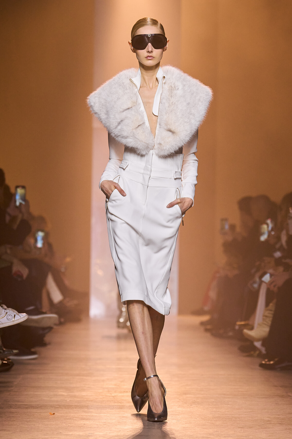 Model walking a runway in a white tailored dress with a large faux fur collar and dark sunglasses.