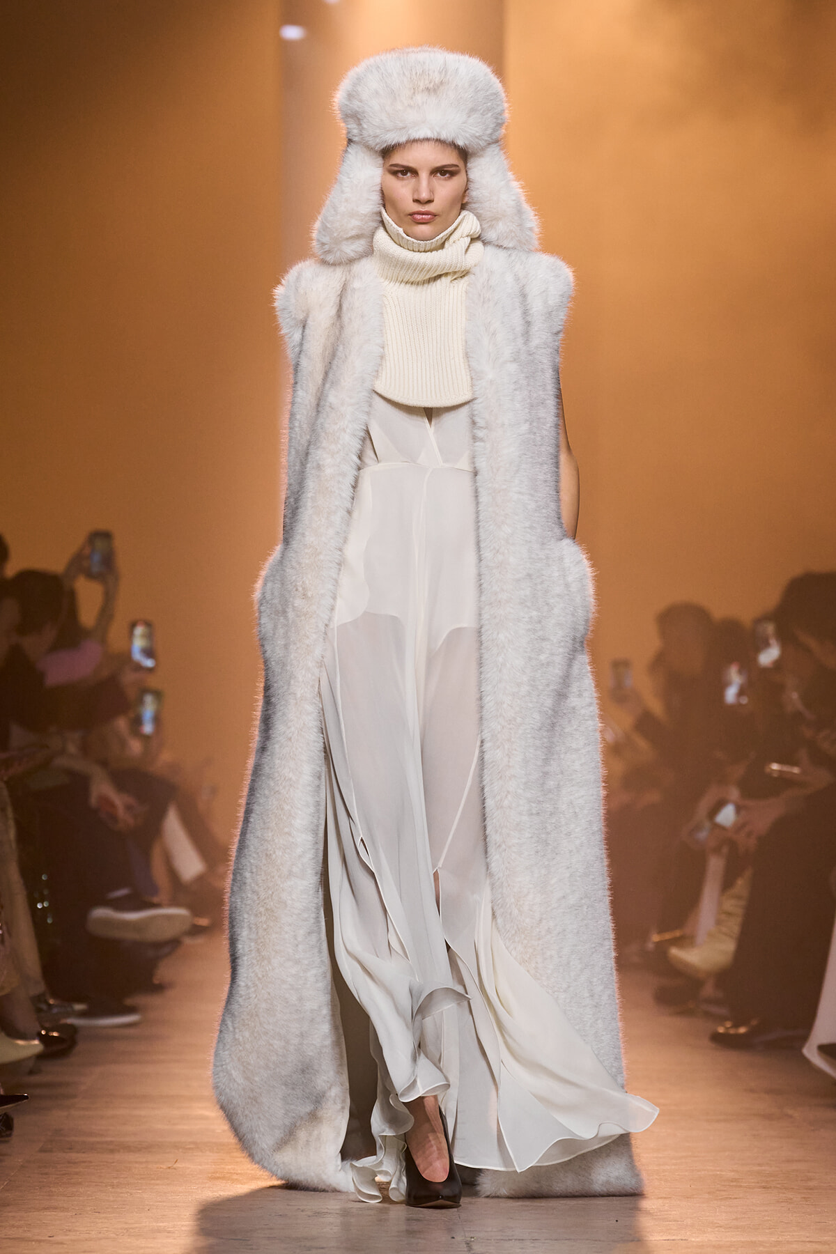 Model walking the runway in a long white fur coat and oversized fur hat, paired with a cream turtleneck and flowing dress step-trailing behind.
