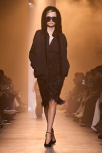 Model walks the runway in a black fur-trim hooded coat, sequined black dress, and oversized sunglasses.