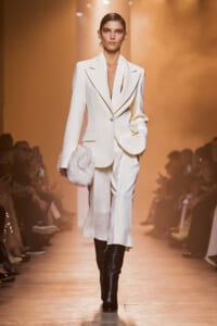 Model on a runway wearing a white tailored suit with a long blazer, wide lapels, and black knee-high boots, carrying a fluffy white fur clutch.