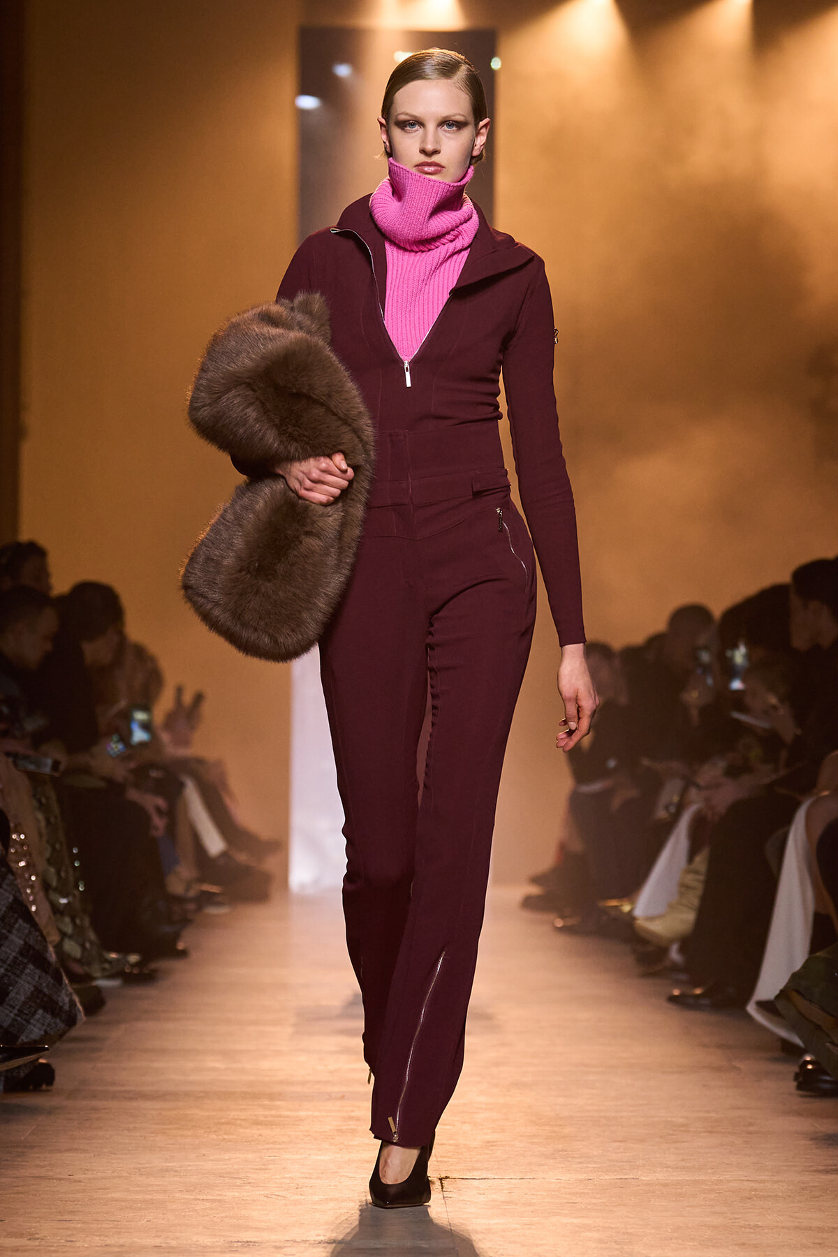 Model walks the runway in a burgundy zip-front jumpsuit paired with a bright pink turtleneck, holding a large fur stole under her arm.