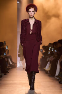 Runway model in a burgundy sequined blazer, matching blouse and wide-leg pants, paired with black knee-high boots, walking the catwalk amid warm lighting.
