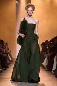 Model on a runway wearing an emerald green strapless gown with a tied waist, carrying a dark fur clutch.