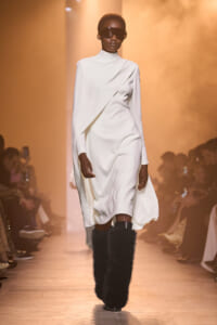 Model walks a runway in a white draped, high-neck dress paired with black furry knee-high boots and oversized sunglasses on a warm-lit catwalk.