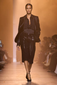 Model walking down an orange-lit runway in a black satin pantsuit with a ruffled blouse and statement shoulders, pairing with black ankle-strap heels.