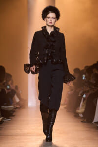 Model on a runway wearing a black textured jacket with glossy ruffles, black pants, knee-high boots, and a black fur hat.