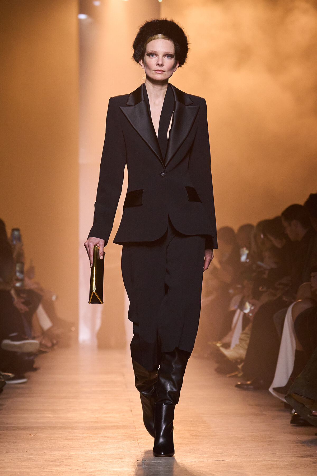 Model walks the runway in a tailored black pantsuit with a deep V-neck, black boots, a black fur hat, and a gold clutch in hand.