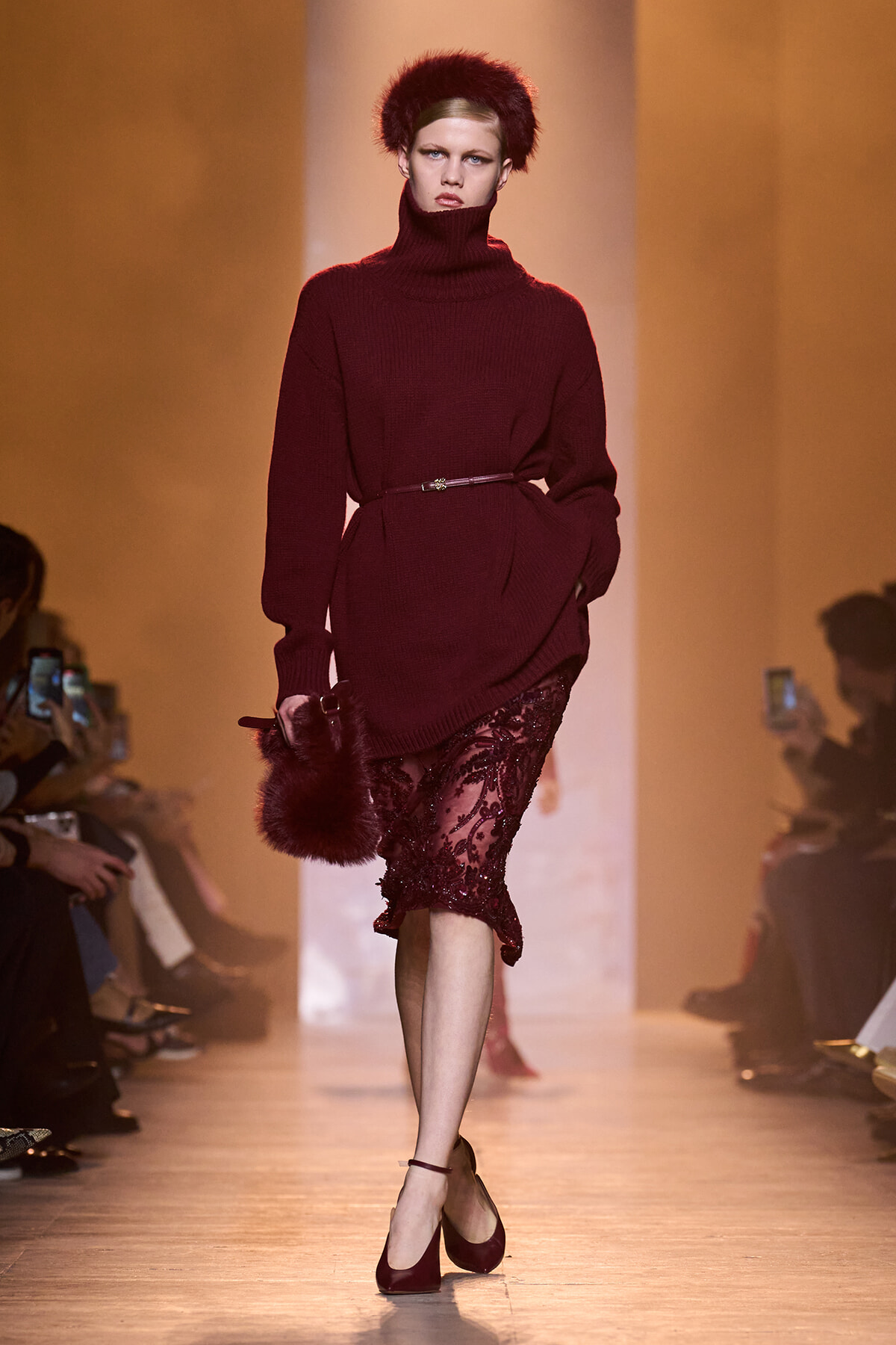 Runway model in a burgundy ensemble: oversized turtleneck sweater belted at the waist, lace midi skirt, burgundy heels, and a fur hat with a fur clutch.