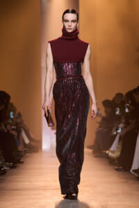 Model on a runway wearing a burgundy turtleneck sleeveless top over a sequined burgundy-black gown with a thin belt, holding a small clutch.