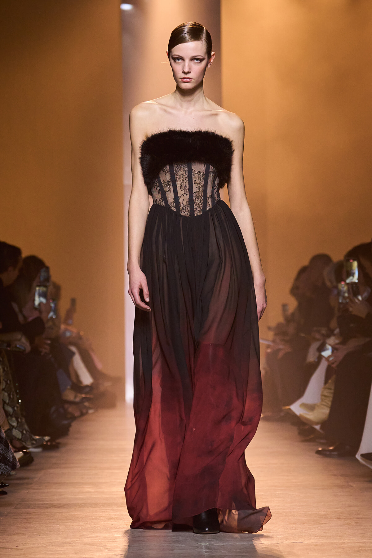 Model walking a runway in a strapless black and burgundy ombre gown with a lace bodice and dark fur trim at the top.