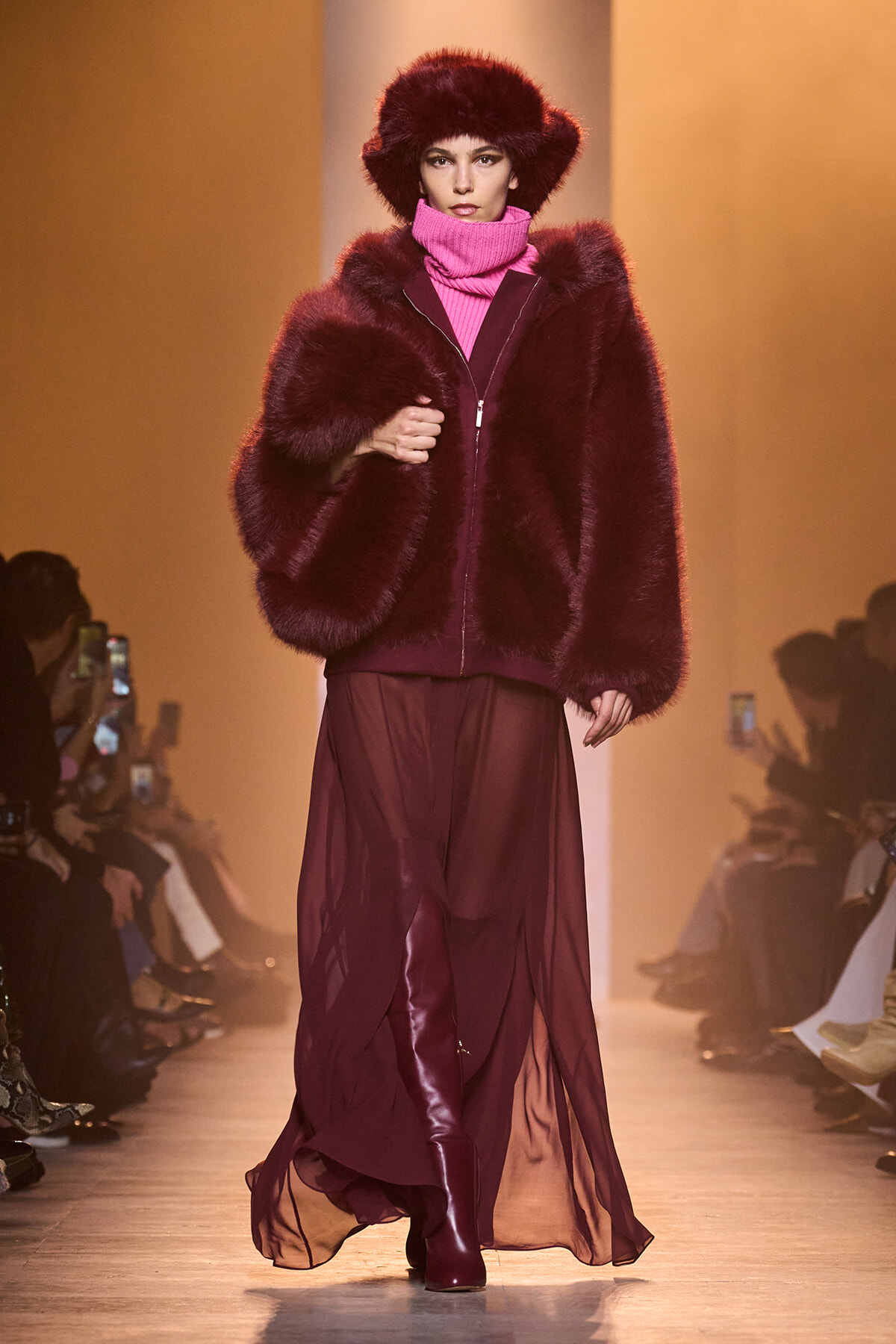 Model on a runway wearing a burgundy fur coat, pink turtleneck, matching hat, and burgundy skirt with knee-high boots; audience in background.